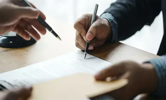 Two hands engaged in a formal signing process, with one hand holding a pen over a document and the other handing over a folder.