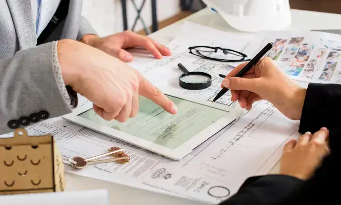 Two professionals discuss a document on a tablet, surrounded by blueprints, glasses, and tools on a table.